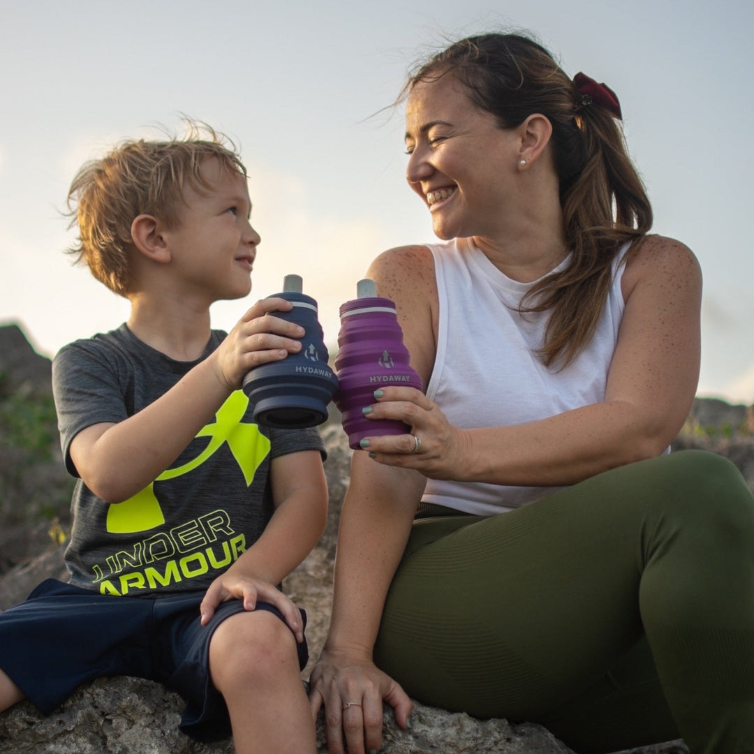 Woman and child sitting on a rock, holding HYDAWAY collapsible water bottles with a scenic background.