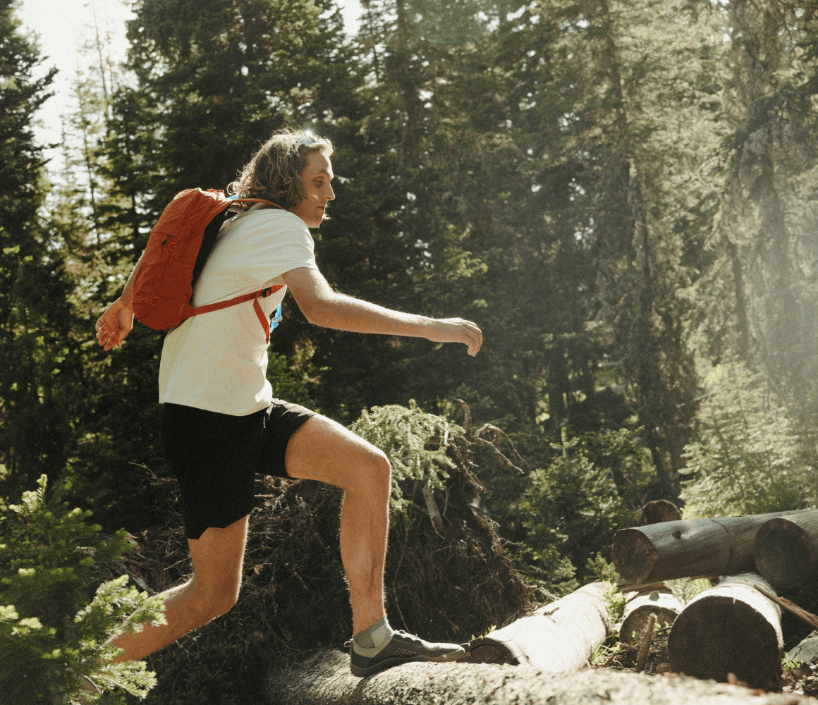 Person hiking through a sunlit forest wearing the Pakt Stash Packable Backpack, a lightweight 16L collapsible daypack designed for travel and outdoor adventures.