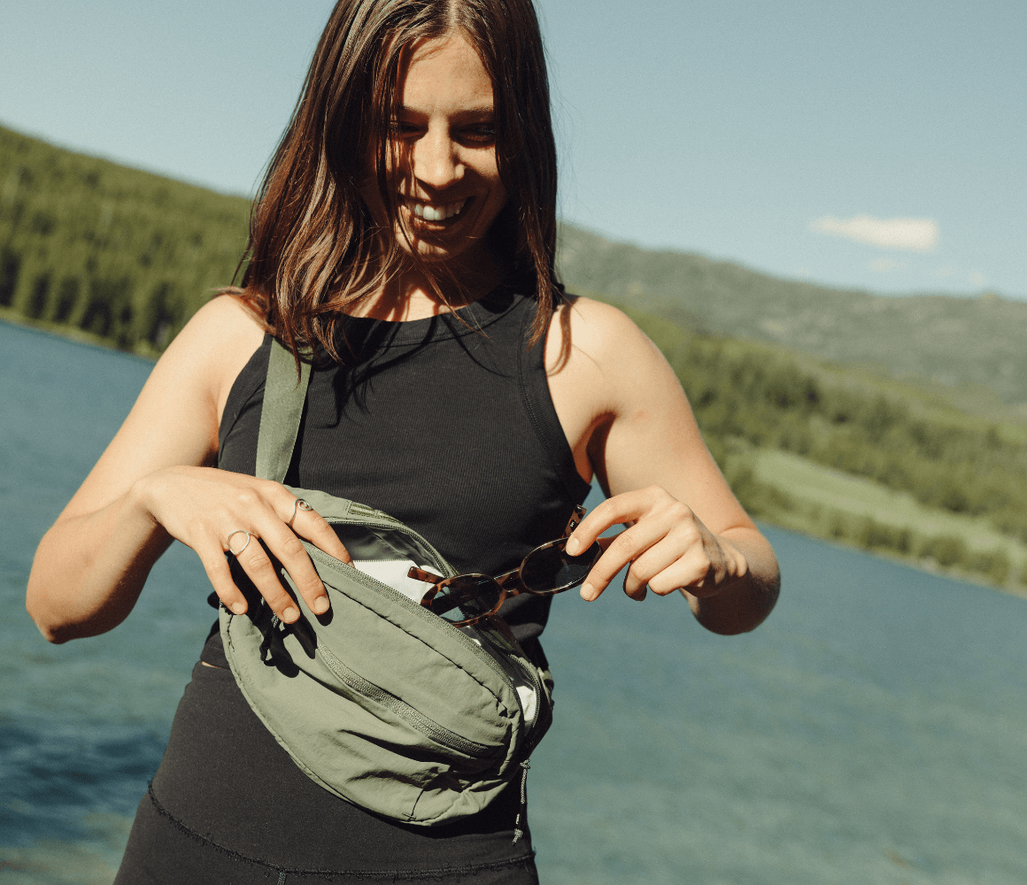 Person opening a Pakt Stash Packable Sling beside a mountain lake, showing the lightweight 4L crossbody bag designed for travel, sightseeing, and everyday adventures.