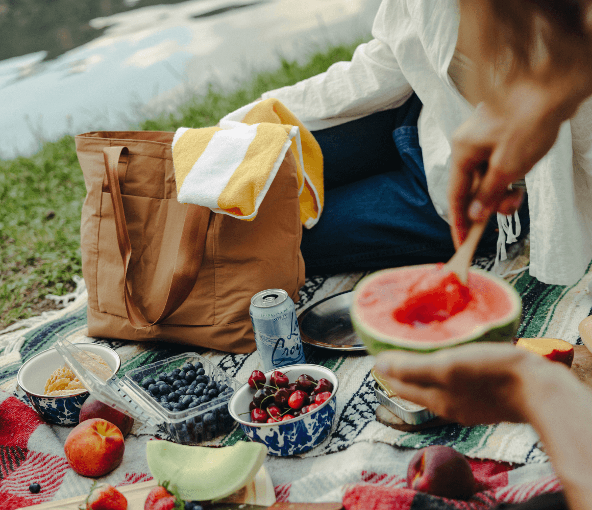 Packable tote bag holding picnic essentials beside a lakeside blanket, showcasing the spacious 20L Pakt Stash Tote for travel, shopping, and outdoor adventures.